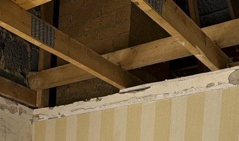 Site photograph looking up into an opened ceiling void, showing timber ceiling joists and a masonry chimney breast above a stripped-back partition wall